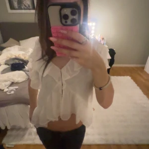 Vit volangblus med knappar - Supersöt vit blus med volanger och knappar framtill. Den är croppad och har korta ärmar med lätt transparent tyg. Perfekt för dig som gillar romantisk och luftig stil. Blusen har v-ringning och volangdetaljer längs både ärmar och nederkant.