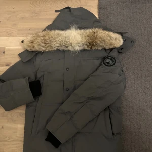 Grå dunjacka Canada Goose med pälskrage - Säljer en grå dunjacka från Canada Goose med stor huva och fluffig beige pälskrage. Jackan har svarta ribbade muddar, diskret logga på ärmen och flera praktiska fickor. Perfekt för kalla vinterdagar och riktigt snygg streetstil.