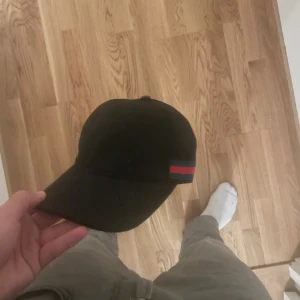 Svart Gucci keps med GG-mönster - Svart keps från Gucci med klassiskt GG-monogram över hela ytan. Kepsen har en böjd skärm och justerbar rem baktill. Sidorna pryds av Guccis ikoniska gröna och röda ränder. Tillverkad i bomullsmix med exklusiv känsla.