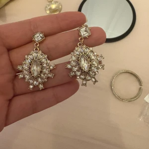 Glittriga örhängen med kristaller - Säljer ett par statement örhängen i guldton med massor av gnistrande klara kristaller. De har en unik, blomliknande form med en stor avlång kristall i mitten och mindre stenar runtom. Perfekta för dig som vill sticka ut och glänsa extra mycket.