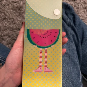 Pennskrin med vattenmelonmotiv - Säljer ett pennskrin med ett lekfullt motiv av en vattenmelon och randiga ben med rosa sneakers. Pennskrinet är ljusgult med blå prickar och har en vit knapp för stängning. Perfekt för dig som vill ha ett färgglatt och unikt fodral till dina pennor med tillbehör. Bilden skiftar sig