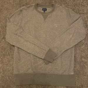 Grå sweatshirt från GANT - Säljer en stilren grå sweatshirt från GANT med broderad logga på bröstet. Tröjan har rund halsringning, ribbade muddar och är tillverkad i mjuk bomullsmix. Hör gärna av er vid frågor eller funderingar