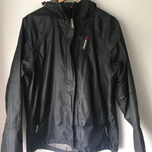 Svart vind och rengjacka - Svart vindjacka från Didrickssons med huva och dragkedja framtill. Jackan har en sportig look, diskret logga på bröstet och är tillverkad i lätt syntetmaterial. Perfekt för blåsiga dagar och enkel att packa ner i väskan.