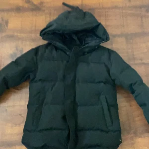 Svart pufferjacka från Canada Goose - Säljer en svart pufferjacka från Canada Goose med huva och klassisk logga på ärmen. Jackan har dragkedja framtill och är vadderad för extra värme. Perfekt för kalla dagar och har en clean, stilren look som funkar till allt.