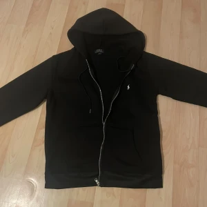 Svart hoodie från Polo Ralph Lauren - Svart hoodie från Polo Ralph Lauren med klassisk vit logga på bröstet. Tröjan har huva med snörning, hel dragkedja och en stor ficka framtill. Materialet är mjuk bomull och passformen är normal. Perfekt för chill dagar och streetwear-stil. Priset kan diskuteras 