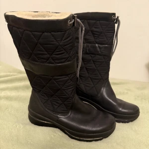 Svarta vinterstövlar  - Svarta stövlar med quiltat mönster och snygga detaljer i skinn. Insidan är fodrad med mjuk ullblandning för extra värme. Stövlarna har dragsko upptill och en robust platt sula, perfekta för kalla dagar.