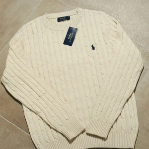 Vit stickad tröja från Polo Ralph Lauren - Snygg vit kabelstickad tröja från Polo Ralph Lauren med klassisk blå broderad logga på bröstet. Tröjan har rund hals, ribbade muddar och är långärmad. Perfekt för dig som gillar stilrena och tidlösa plagg med premiumkänsla.