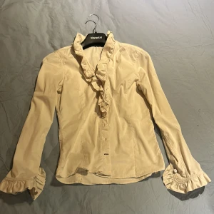 Beige volangskjorta  - Supersöt beige skjorta med volangdetaljer längs kragen och ärmsluten. Långärmad modell i mjuk bomull, knäppning framtill och figurnära passform. Perfekt för dig som gillar romantiska vibes och snygga detaljer. Mycket mer benvit i verkligheten!💕fått av mamma så en riktigt fin vintage tröja!