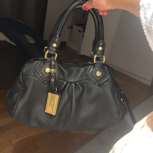 Svart handväska från Marc by Marc Jacobs - Snygg svart handväska i skinn från Marc by Marc Jacobs med guldfärgade metalldetaljer och dubbla handtag. Väskan har en mjuk, rundad form och en dekorativ metallbricka framtill. Perfekt för dig som gillar klassisk och stilren design. Pris kan diskuteras 