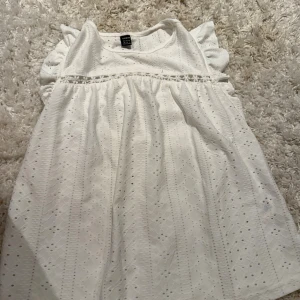 Vit somrig blus med volangärm - Supersöt vit blus med broderade detaljer och små hålmönster. Volangärmar och en söt spetskant under bysten ger en romantisk känsla. Perfekt för varma dagar och midsommarfirande.