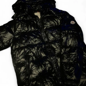 Svart glansig dunjacka från Moncler - Säljer en svart glansig dunjacka från Moncler med huva och dragkedja framtill. Jackan har quiltad design, två fickor med dragkedja och Moncler-logga på ärmen. Perfekt för kalla dagar och riktigt snygg streetstil.