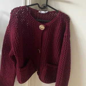 Vinröd stickad kofta från Zara - Säljer en vinröd stickad kofta från Zara med grovstickat mönster, två fickor framtill och snygga guldfärgade knappar.
