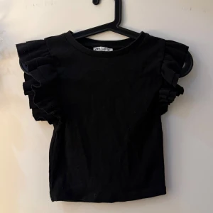 Svart ribbad topp med volangärmar Zara - Svart ribbad topp från Zara i storlek S. Toppen har korta ärmar med breda volanger som ger en cool och trendig look. Rund halsringning och croppad passform. Materialet är mjukt och stretchigt, perfekt för att lyfta din outfit.
