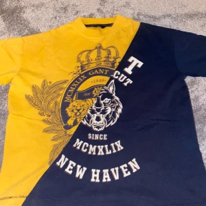 Gul och marinblå t-shirt från GANT - Unik t-shirt från GANT i gul och marinblå färg med coolt tryck av krona, tiger och texten 'NEW HAVEN'. Snyggt delad design med grafiska detaljer och college-vibe. Rund hals och korta ärmar, tillverkad i mjuk bomull. Perfekt för dig som gillar statement-plagg.