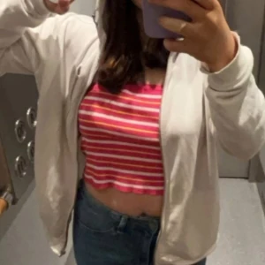 Rosa, vit och gul randig croppad linnetopp - Säljer en färgglad croppad linnetopp med rosa och gula ränder. Toppen är ärmlös och har en vågig kant nedtill som ger en cool touch. Perfekt för varma dagar och att styla med jeans eller shorts.