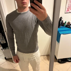 Grå merinoulltröja från Riley - Snygg grå tröja från Riley i mjuk merinoull. Rund halsringning och långärmad modell med ribbade muddar vid ärmslut och nederkant. Perfekt för dig som gillar stilrena och klassiska plagg med skön passform. Sitter som xs/xxs