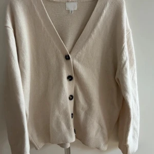 Beige kofta från H&M med knappar - Snygg beige kofta från H&M i storlek M. Koftan har v-ringning, svarta knappar framtill och är långärmad. Materialet är en mix av polyester, akryl och ull som gör den mjuk och skön. Perfekt att slänga på sig när det är lite kyligt ute.