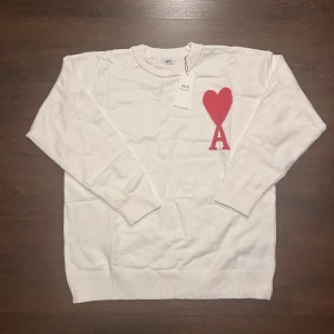 Vit stickad tröja från Ami storlek M - Helt ny vit stickad tröja från Ami Paris med röd hjärt- och A-logga på bröstet. Rund halsringning och ribbade muddar vid ärmslut och nederkant. Perfekt för dig som gillar stilrena detaljer och vill ha något som sticker ut lite extra.