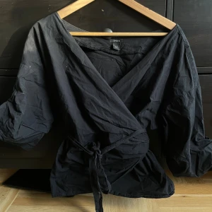 Svart omlottblus från Lindex - Svart omlottblus från Lindex med knyt i midjan och djup v-ringning. Blusen har trekvartsärmar och är tillverkad i mjuk bomull. Perfekt för dig som gillar stilrena och enkla plagg med en cool twist.