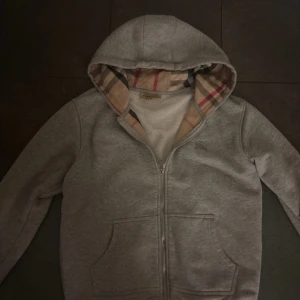 Grå Burberry hoodie med dragkedja - Snygg grå hoodie från Burberry med klassiskt rutigt foder i huvan och broderad logga på bröstet. Tröjan har dragkedja framtill, två fickor och är tillverkad i mjuk bomull. Perfekt för dig som vill ha en stilren och lyxig vardagslook.