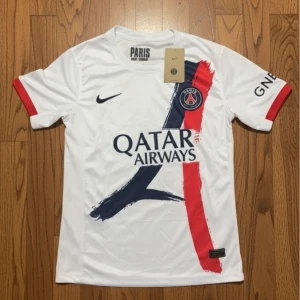 PSG vit fotbollströja Nike - Snygg vit PSG fotbollströja från Nike med tryck av Qatar Airways på bröstet. Tröjan har korta ärmar med röda detaljer och ett grafiskt mönster i marinblått och rött framtill. Materialet är lätt och andas, perfekt för match eller träning.