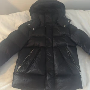 Svart pufferjacka från BOSS med huva - Snygg svart pufferjacka från BOSS med avtagbar huva, diskret mönstrad yta och silverfärgade dragkedjor. Jackan har stora fickor med dragkedja och tryckknappar framtill. Perfekt för kalla dagar och ger en cool vibe till din outfit.