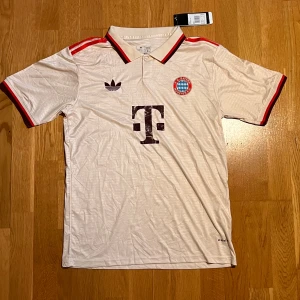 Bayern München vit Adidas fotbollströja - Snygg vit Bayern München fotbollströja från Adidas med klassisk krage och röda detaljer på axlar och ärmslut. Broderat klubbmärke på bröstet, Adidas-logga och stor T-sponsor framtill. Tillverkad i lätt polyester med Aeroready-teknologi. Har legat hemma ett tag pga att den var för att den var för stor och har ej orkat sälja. Kan sänka pris vid snabb affär!
