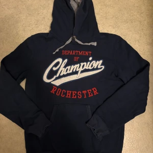 Mörkblå Champion hoodie med tryck - Mörkblå hoodie från Champion med stor broderad logga och texten 'Department of Champion Rochester' i vitt och rött framtill. Tröjan har huva med grått foder och snörning samt en klassisk känguruficka. Perfekt för en sportig och avslappnad stil.