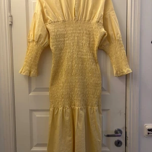 Ljusgul långklänning från Gina Tricot - Supersöt ljusgul långklänning från Gina Tricot med smockad midja och ärmar, samt en volang nedtill. Klänningen har rund halsringning och knäppning bak i nacken. Tillverkad i mjuk bomull, perfekt för sommarens alla tillfällen.