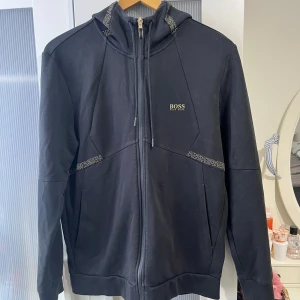Hugo Boss Hoodie / kofta / luvtröja med zip - En mycket fin Hugo Boss huvtröja med dragkedja/ kofta med zip Svart färg med guldiga detaljer  Storlek M på lappen I nyskick Skicka meddelande vid några funderingar Kolla gärna även in mina andra annonser