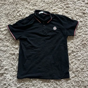 Svart Moncler pikétröja med röd kant - Svart pikétröja från Moncler med klassisk krage och knappar framtill. Tröjan har röda och vita detaljer på krage och ärmslut samt Moncler-logga på bröstet. Perfekt för dig som gillar stilrena och sportiga plagg.