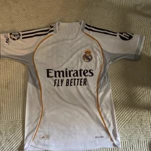 Real Madrid T-shirt nmr 7 vin jr - Säljer en officiell Real Madrid fotbollströja från Adidas med Vini Jr. och nummer 7 på ryggen. Tröjan är vit med svarta detaljer, guldiga linjer och klubbmärke på bröstet. Kortärmad modell i funktionsmaterial med HEAT.RDY-teknologi och tryckta sponsorer.