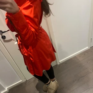 Röd trenchcoat med bälte och dragkedja - Snygg röd trenchcoat med midjebälte och dragkedja framtill. Jackan uttrycker kurvor och är elegant