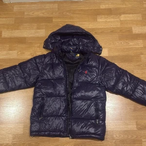 Mörkblå pufferjacka Polo Ralph Lauren - Säljer en mörkblå pufferjacka från Polo Ralph Lauren med huva och dragkedja. Jackan har två fickor med dragkedja och klassisk röd logga på bröstet. Materialet är glansigt och känns lätt men ändå varmt, perfekt för kyliga dagar.