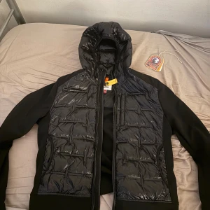 Svart hybridjacka Parajumpers XL - Svart hybridjacka från Parajumpers i modellen Techno Hybrid. Jackan har quiltad, glansig front och huva, samt släta svarta ärmar och sidor. Märkespatch på ärmen och dragkedja framtill. Perfekt för dig som vill ha en sportig och modern look. Pris kan diskuteras!