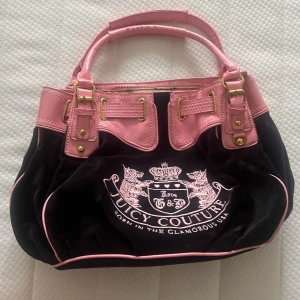 Juicy Couture svart och rosa handväska - Juicy Couture handväska i svart sammet med rosa detaljer i skinn och guldiga metalldetaljer. Väskan har broderad logga framtill, dubbla handtag och innerfoder med kronmönster. Utrustad med dragkedja och hjärtformad nyckelring i guld. Perfekt för dig som gillar statement-accessoarer.