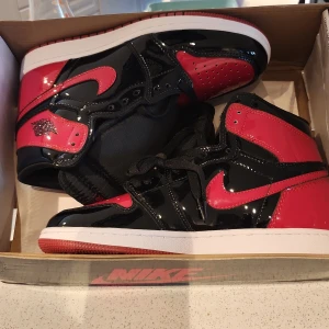 Nike Air Jordan 1 Patent Bred 6.5Y - Säljer ett par Nike Air Jordan 1 Patent Bred i storlek 6.5Y. Skorna har en blank finish i svart och rött med klassisk vit sula och röda detaljer. Ikonisk Jordan-logga på sidan och extra röda skosnören medföljer. Perfekta för dig som vill sticka ut med en tidlös sneaker.