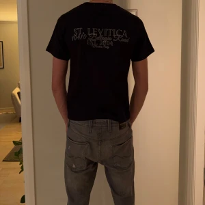 Svart t-shirt från Saint Levitica - Svart t-shirt från Saint Levitica med vit ST-logga på bröstet och stor text på ryggen. Klassisk passform med rund hals och korta ärmar. Skön och stilren, perfekt till jeans eller shorts.