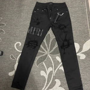 Svarta skinny jeans från Amiri, strl 30 - Svarta skinny jeans från Amiri med slitningar och trasiga detaljer på benen. Stora AMIRI-bokstäver i läder på vänster lår och ett A på baksidan. Tillverkade i bomull med lite elastan för stretch. Snygg streetstil med smal passform.