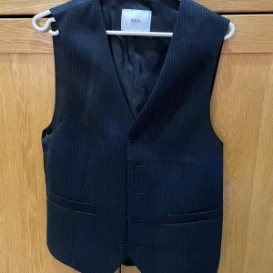 Svart randig kostymväst från Bläck - Snygg svart kostymväst från Bläck med tunna vita ränder. Västen har klassisk v-ringning, tre knappar framtill och två fickor. Materialet är slätt och känns dressat, perfekt för att lyfta din outfit. Passformen är normal och designen är stilren.