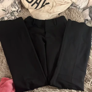 Svarta högmidjiga kostymbyxor från BikBok - Snygga svarta kostymbyxor från BikBok i storlek 34. Byxorna har klassisk rak passform med hällor för bälte och stängs framtill. Perfekta för en clean och stilren look, tillverkade i ett mjukt och följsamt material.