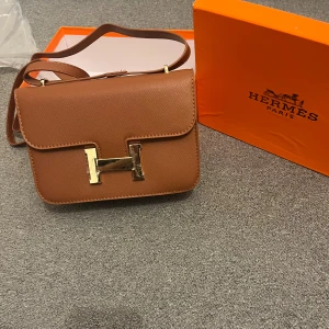  brun axelväska i skinn - Säljer en lyxig brun axelväska  med ikoniskt  i guldmetall. Väskan är tillverkad i exklusivt skinn och har en stilren, rektangulär form med justerbar axelrem. Perfekt för dig som vill ha något klassiskt och tidlöst med en touch av Paris.