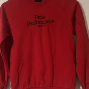 Röd Peak Performance sweatshirt - Röd sweatshirt från Peak Performance med svart broderad logga på bröstet. Klassisk rund halsringning och långa ärmar. Perfekt för dig som gillar en enkel men snygg stil.