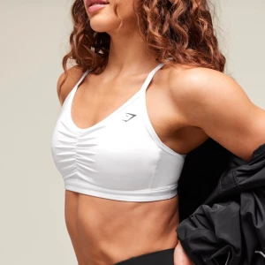 Vit sportbh från Gymshark - Säljer en vit sportbh från Gymshark med smala korsade axelband och snygg rynkdetalj framtill. Toppen har en sportig vibe och är tillverkad i stretchigt syntetmaterial som passar perfekt för träning. Diskret Gymshark-logga på bröstet. Ingen vaddering. Använd ett fåtal gånger 🥰