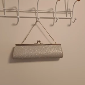 Glittrig silver kuvertväska med kedja - Snygg kuvertväska i silver med glittrig yta och metallkedja. Väskan har ett klassiskt metallspänne upptill och en stilren, avlång form. Perfekt för att lyfta din outfit med lite extra bling.