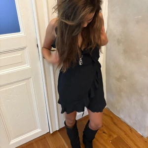 Svart omlottklänning med volang - Säljer en svart kort omlottklänning med volangkant och offshoulder-design. Klänningen har en snygg knytning i midjan och är tillverkad i ett lätt, mjukt material som ger en skön känsla. Perfekt för dig som gillar stilrena och trendiga plagg.