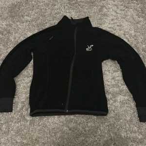 Svart fleecejacka RevolutionRace 152 - Svart fleecejacka från RevolutionRace i ungdomsstorlek 152. Jackan har hel dragkedja, hög krage och logga på bröstet och ryggen. Materialet är mjukt och värmande fleece, perfekt för kyliga dagar. Stilren design med lång ärm och diskreta detaljer.