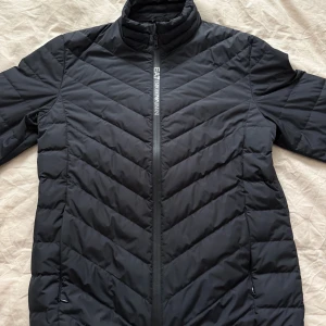 Svart pufferjacka från Emporio Armani - Svart pufferjacka från Emporio Armani EA7 med diskret logga på bröstet och dragkedja framtill. Jackan har ståkrage, horisontella och snedställda quiltade sömmar samt två fickor med dragkedja. Materialet är syntet och jackan har en clean, sportig vibe. Nypris 2500kr