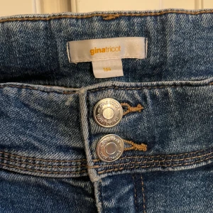 Blå bootcut jeans från Gina Tricot - Snygga blå jeans från Gina Tricot i storlek 164 med klassisk bootcut passform. Jeansen har två knappar fram, fem fickor och coola detaljer med knappar på bakfickorna.