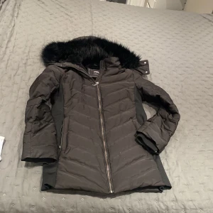 Svart pufferjacka med fuskpäls från Zara - Svart pufferjacka från Zara med snygg fuskpäls på huvan och mjukt pälsfoder inuti. Jackan har dragkedja framtill, quiltad design och lång ärm. Perfekt för kalla dagar och har en stilren look som funkar till många outfits.